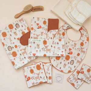 Ensemble de naissance à motif forêt sur fond beige uni, mis en scène avec des accessoires de puériculture. On y voit un protège-carnet, un bavoir, une pochette nomade , des lingettes lavables, une brosse et un peigne en bois, des langes blancs pliés et un petit bracelet pour bébé.