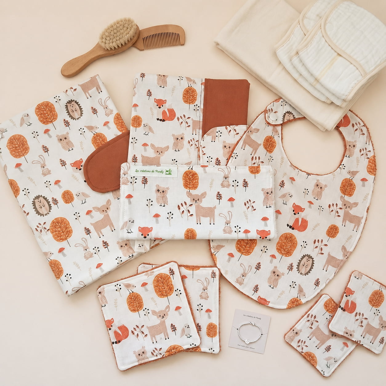 Ensemble de naissance à motif forêt sur fond beige uni, mis en scène avec des accessoires de puériculture. On y voit un protège-carnet, un bavoir, une pochette nomade , des lingettes lavables, une brosse et un peigne en bois, des langes blancs pliés et un petit bracelet pour bébé.