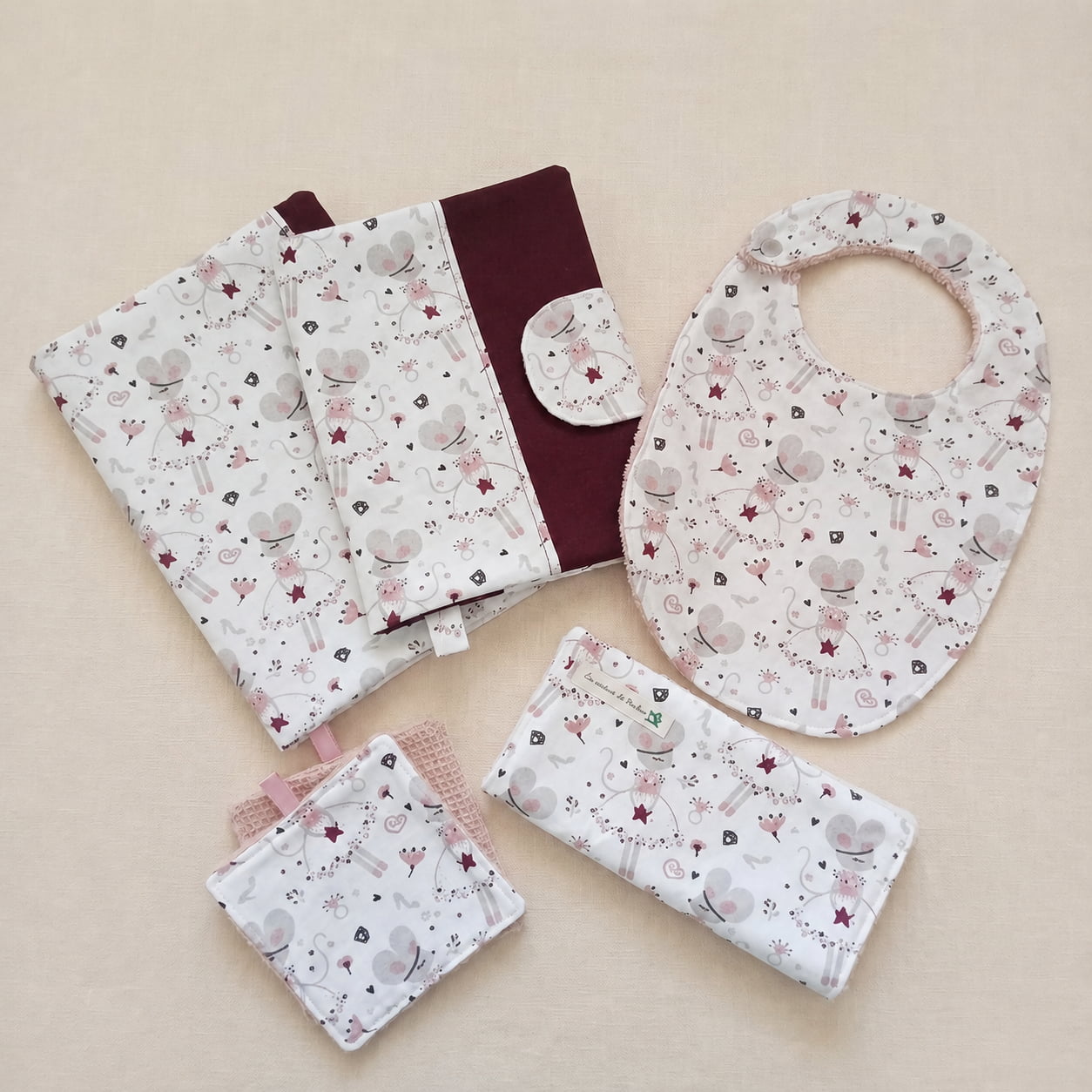 Un ensemble d'accessoires de naissance assortis pour bébé avec un motif de souris ballerine blanche et rose et du tissu bordeaux. L'ensemble comprend un protège-carnet de santé, un bavoir, une pochette à lingettes fermée et des carrés de coton lavables.