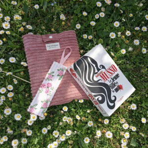 pochette-livre-velours-rose-lecture-jardin-fleurs Pochette de livre en velours rose poudré et son marque-page fleuri posés dans une pelouse de marguerites à côté d'un roman.