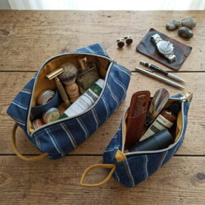 Ensemble de deux trousses de toilette homme en denim rayé. A l'intérieur de chacune d'elles des produits de soin. Posées sur une table en bois vintage